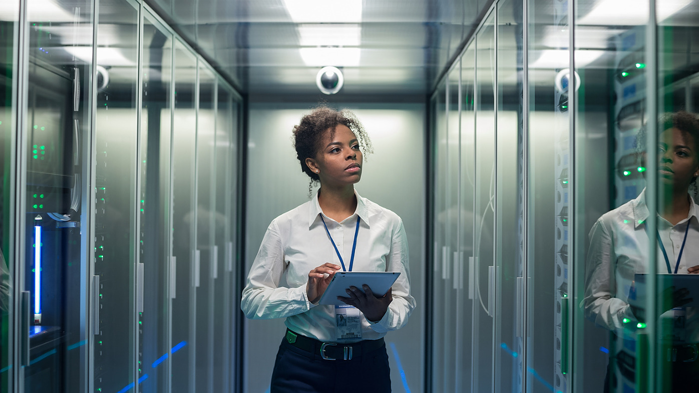 African American woman using tablet while walking in corridor of data center and checking hardware on server racks