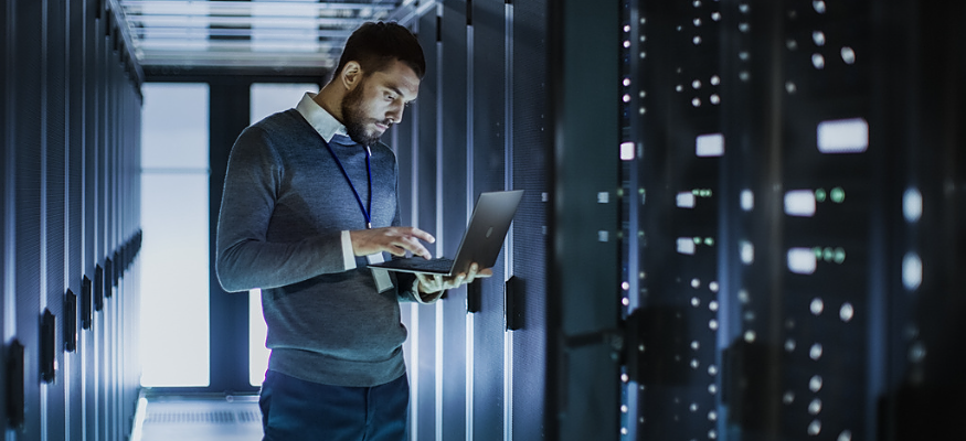 IT Technician Works on Laptop next to a Server Cabinet in Big Data Center. He Runs Diagnostics and Maintenance, Sets System Up.